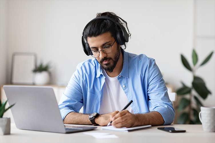 Image shows a young man with dark hair, a beard, and glasses, taking notes in front of an open laptop.